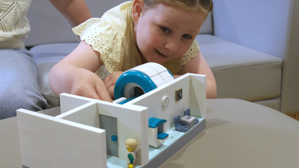 A young girl plays with a LEGO MRI scanner model, learning about medical scans through play.